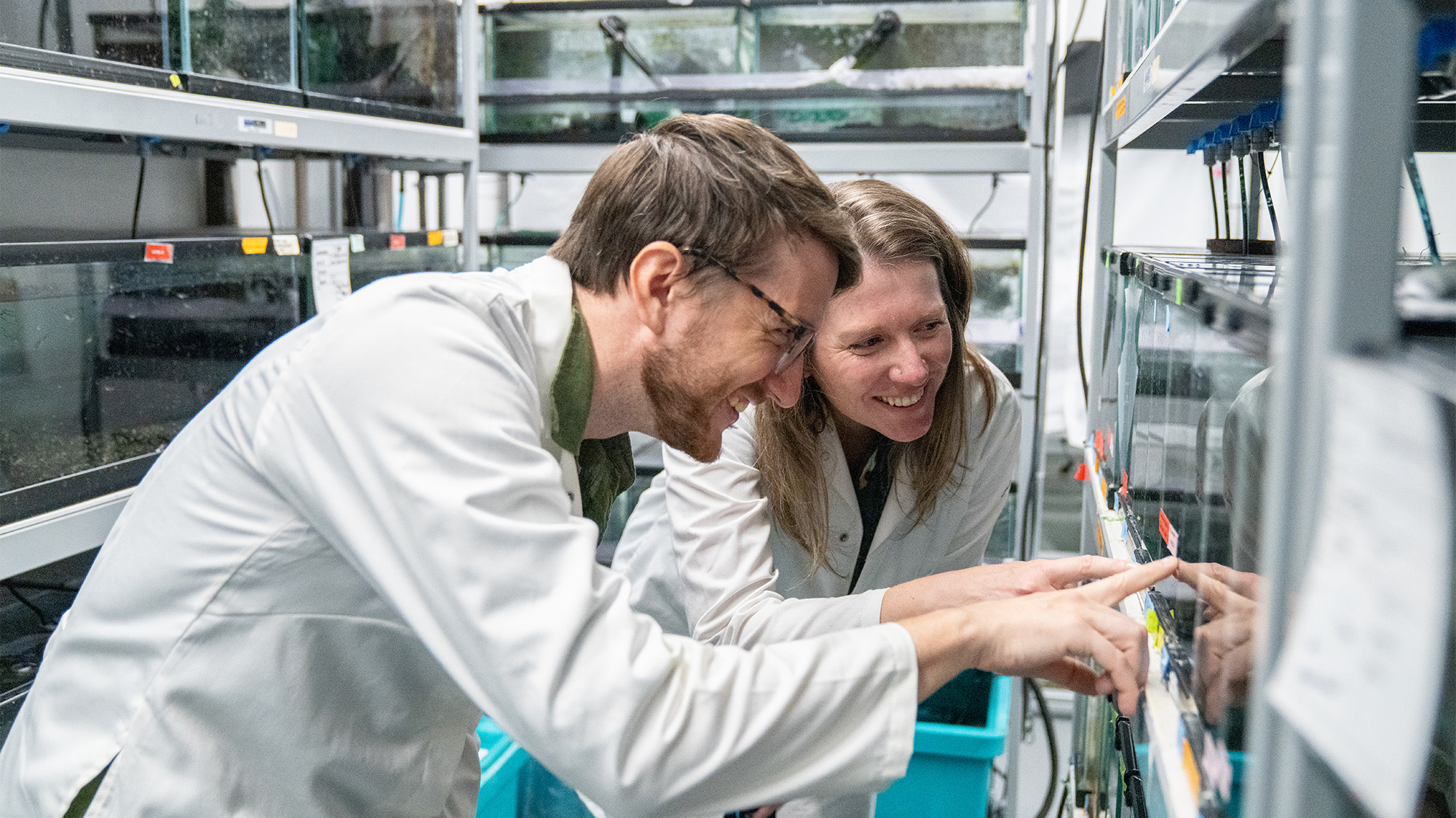 Two scientists in lab coats smiling as they adjust laboratory equipment