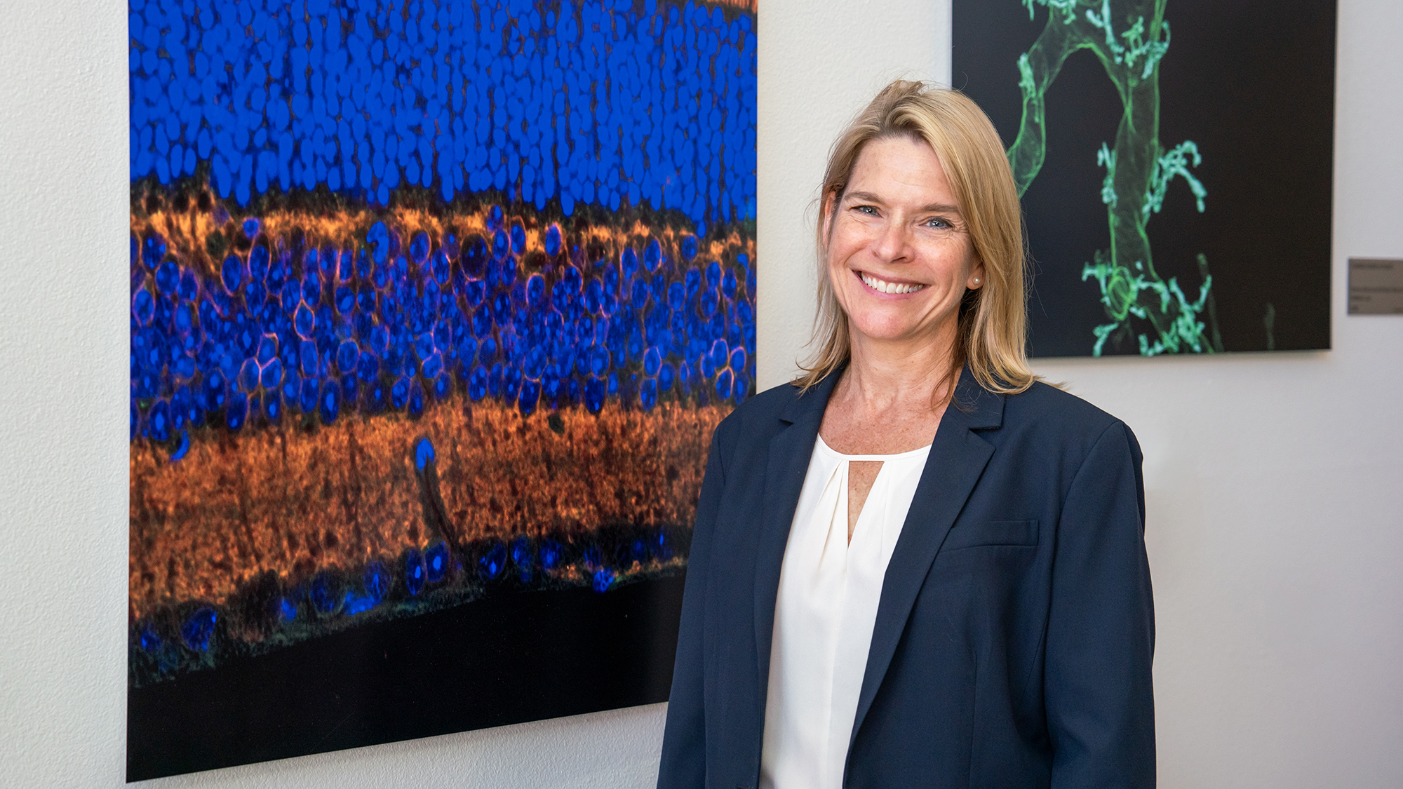 Smiling woman in blazer stands beside colorful microscopic image artwork displayed on a wall.