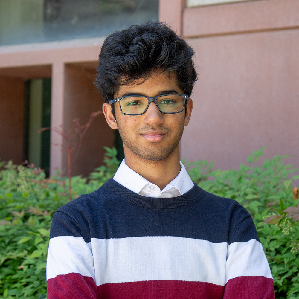 Young man with glasses in striped sweater, slight smile, leafy background