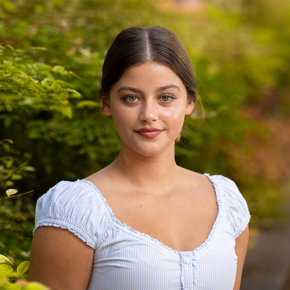 Outdoor portrait of a young woman with dark hair in light blue striped blouse, green foliage background