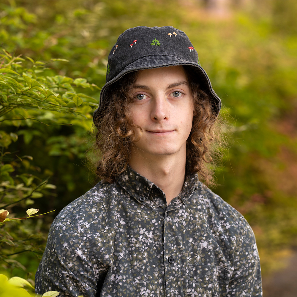 Outdoor portrait photo of young person with curly hair, patterned shirt, black bucket hat