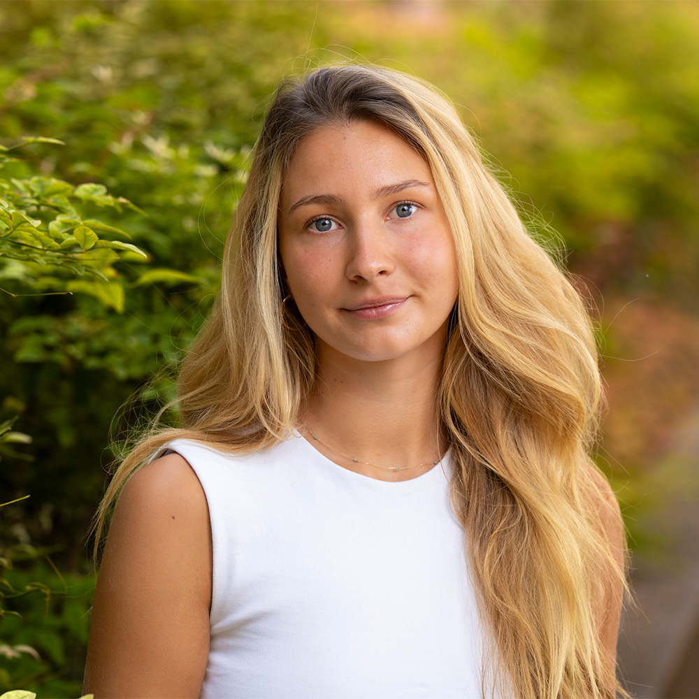 Young woman with long blonde hair in white sleeveless top, soft smile, green foliage background.