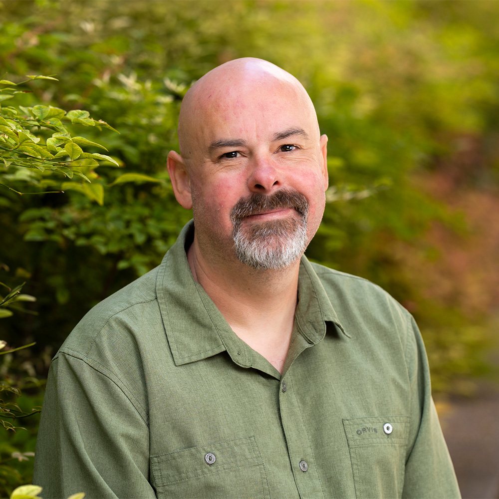 Headshot photo of smiling bald man with goatee wearing green shirt, leafy outdoor background
