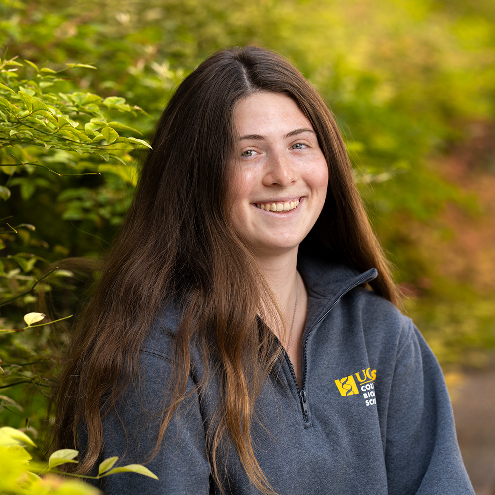 Portrait of smiling young woman with long dark hair in blue fleece, green leafy background