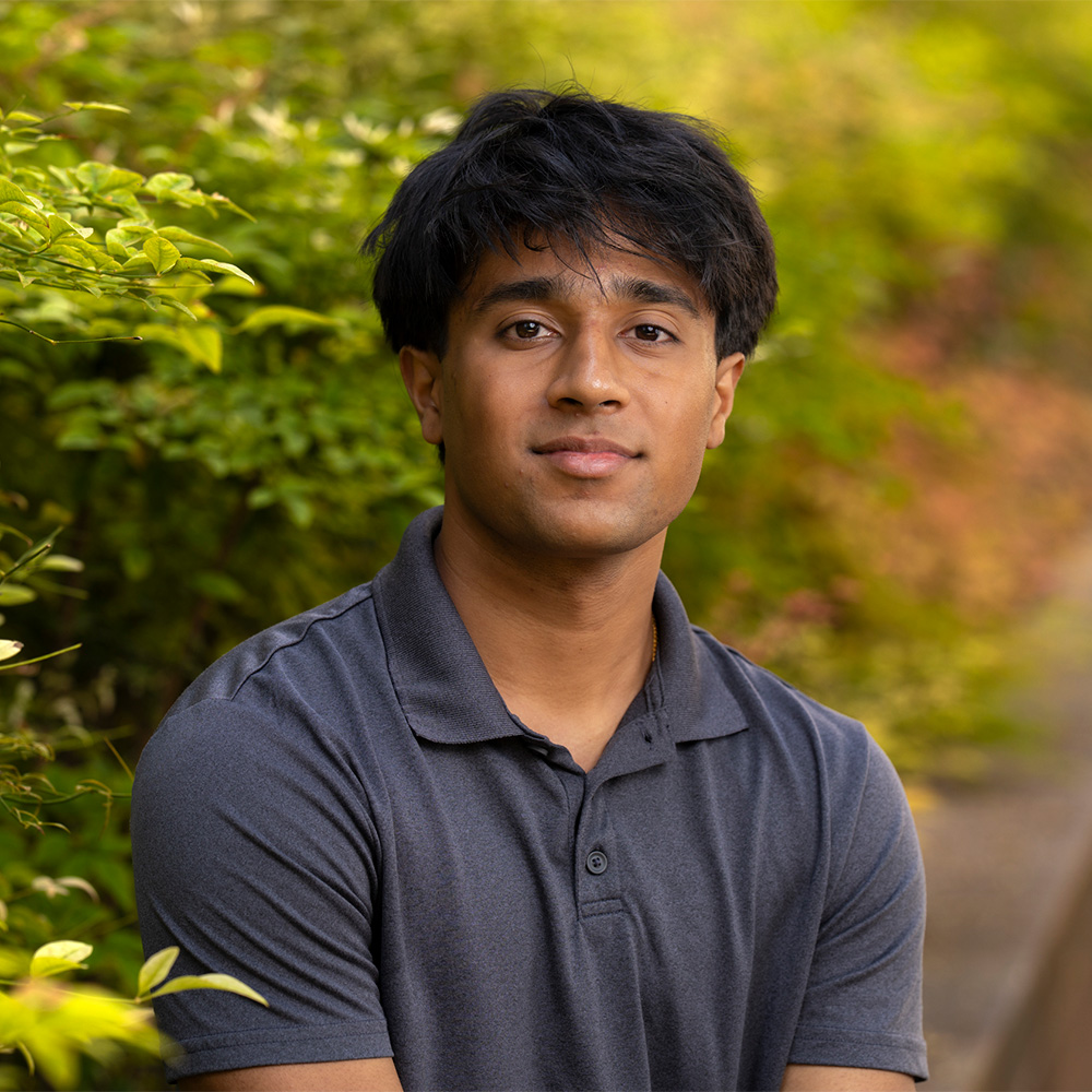 Young man in gray polo seated outdoors, neutral expression, green foliage background, photo