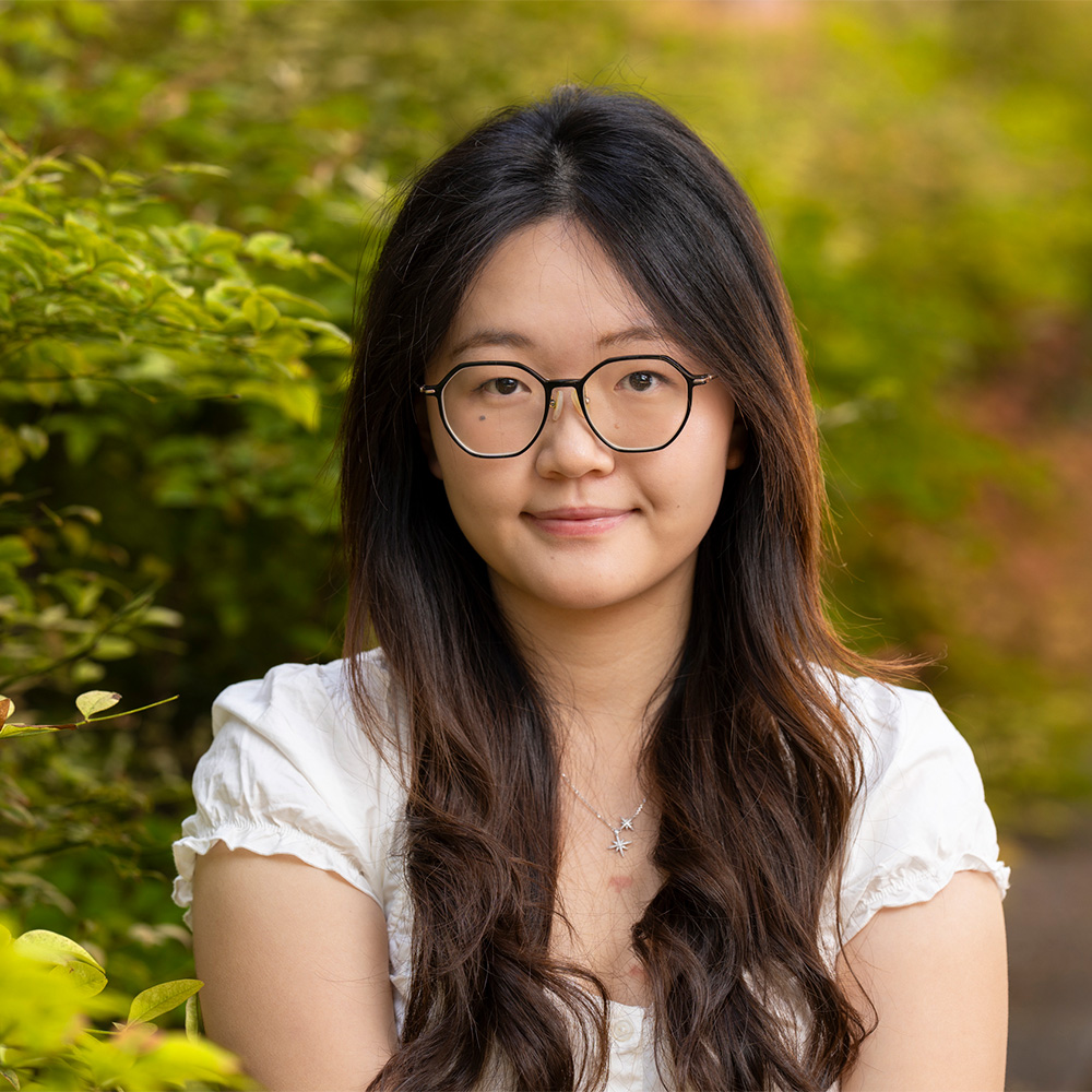 Young woman with long dark hair, glasses, smiling in a green outdoor headshot photo
