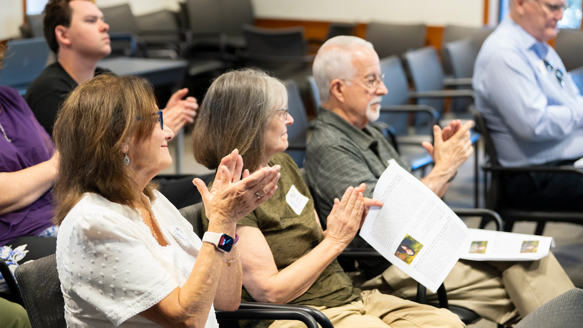Mixed-age audience seated in conference room, clapping; man holds a printed program