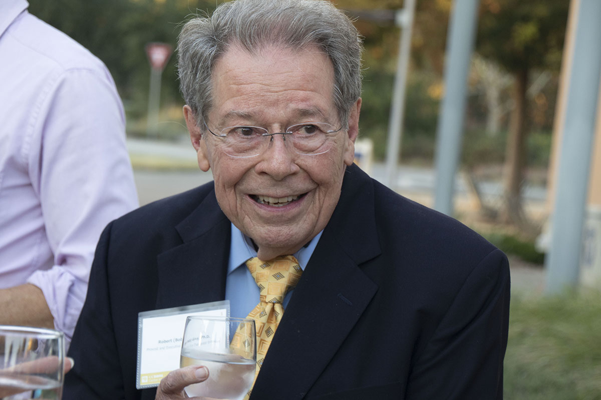Smiling older man in a suit holding a glass, outdoors at a gathering.