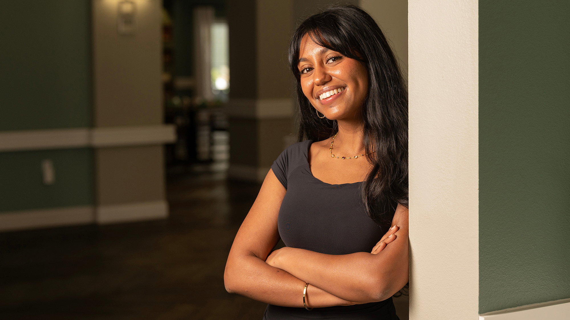A UC Davis student smiles with arms crossed while leaning against an indoor hallway pillar. She has long dark hair, wears a black top and gold jewelry, and stands confidently in soft lighting.