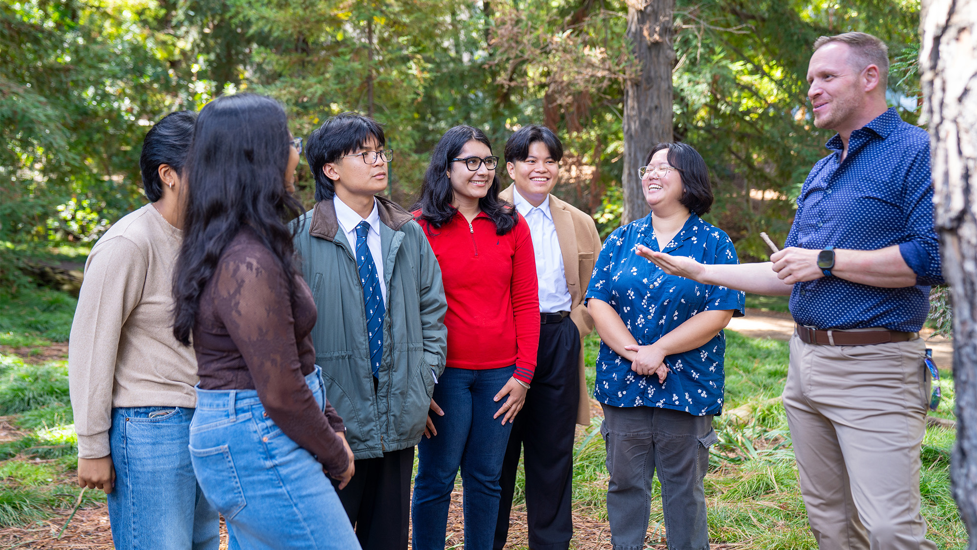 Adults standing outdoors under trees, attentively listening as a man speaks