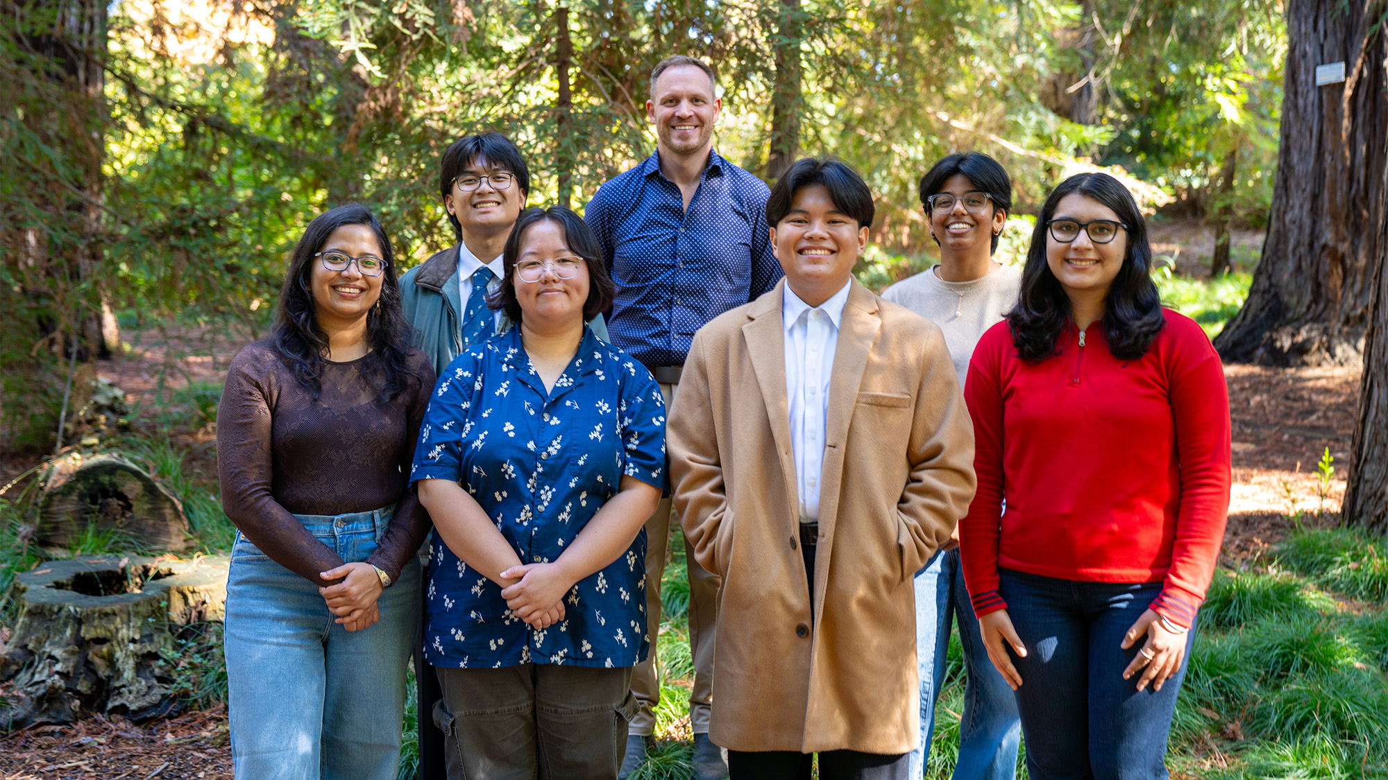 Smiling group standing outdoors among tall trees, casual clothing