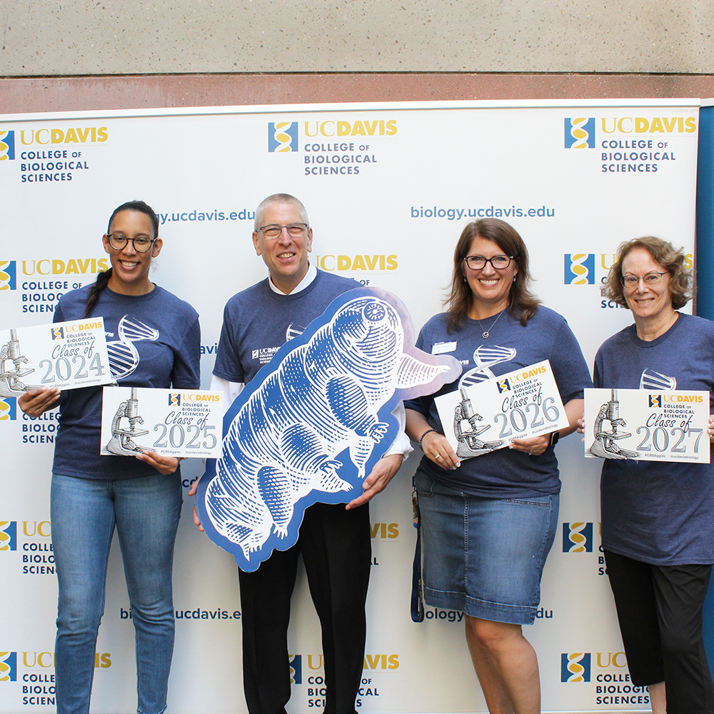 Four smiling individuals wearing UC Davis College of Biological Sciences shirts pose in front of a branded backdrop, holding signs for the Classes of 2024, 2025, 2026, and 2027, along with a large cutout of a tardigrade.