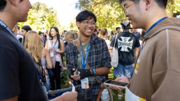 Smiling student wearing glasses chats with two others during an outdoor campus orientation, surrounded by a crowd of peers.