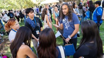 Faculty member with long curly hair speaks to a group of students in a grassy outdoor area during UC Davis orientation.
