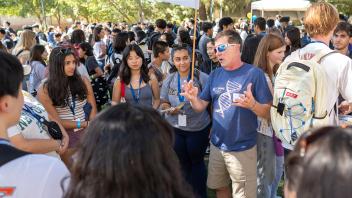Faculty member in sunglasses and a DNA shirt gesture while speaking to a circle of students in a crowded outdoor setting.