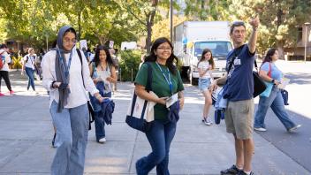 Orientation leader points while walking with students carrying tote bags and shirts across a shaded campus walkway.