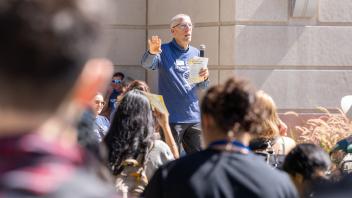 Speaker in blue shirt addresses a crowd outdoors, holding a microphone and orientation flyer in front of a campus building.
