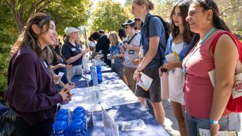Students speak with staff at an outdoor resource fair table lined with flyers, water bottles, and informational materials.