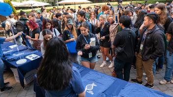 At an outdoor welcome gathering, students wait in line to pick up free blue T-shirts featuring a DNA strand design.