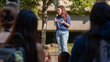 Student in a navy UC Davis shirt speaks into a microphone on an outdoor stage, addressing a crowd under sunny trees.