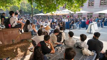 Large group of students gathered in an outdoor courtyard, listening to a speaker under tree shade and patio umbrellas.