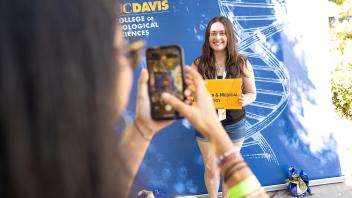 Student poses holding a “UC Davis College of Biological Sciences” sign while another takes her photo against a blue backdrop.