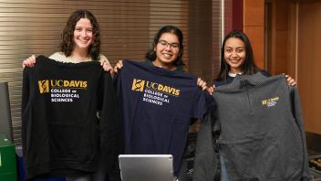 Three smiling young women stand behind a table displaying UC Davis College of Biological Sciences merchandise, holding up sweatshirts and T-shirts with the college logo. They are indoors, and the table in front of them is covered with neatly folded apparel.