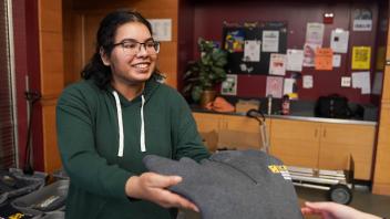 A smiling young woman wearing a green hoodie hands over a folded gray UC Davis College of Biological Sciences sweatshirt to someone off-camera, standing in a room with tables and bulletin boards in the background.
