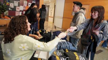 A woman in a floral sweater hands cash to a young woman standing behind a table displaying UC Davis College of Biological Sciences clothing items, as other students wait in line.