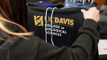 A person holds up a black UC Davis College of Biological Sciences hoodie, showcasing the college's logo and name in white and gold against the dark fabric.