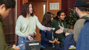 A young woman in a green hoodie and another in a floral sweater assist students purchasing UC Davis College of Biological Sciences merchandise at a table, with various items displayed.