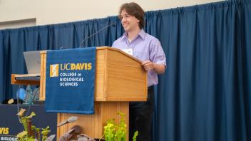 Student in purple shirt speaking at UC Davis awards podium with plants in foreground and College banner hanging on lectern.