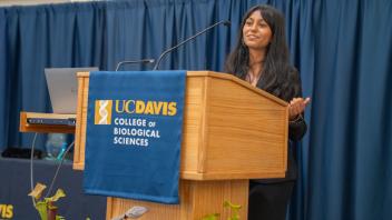 Female student at UC Davis awards ceremony speaks confidently at podium. Decorative pitcher plants and blue curtain provide backdrop.