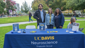 Three people stand behind a UC Davis Neuroscience Graduate Group table outdoors during the Grad Student Fair.