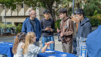 A group of students talk outdoors at the UC Davis Graduate Student Fair, surrounded by tables and trees.