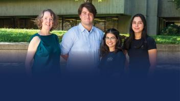 Four individuals stand side by side in front of a building with green foliage. From left to right, the first person is wearing a teal top, the second person is in a light blue shirt, the third person is wearing a dark top with a logo on it, and the fourth person is dressed in black. The setting appears to be outdoors during daylight with ample sunlight casting shadows on the group.