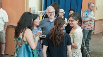 Group of adults mingle and laugh while holding drinks and food at UC Davis event with informational signage in background.