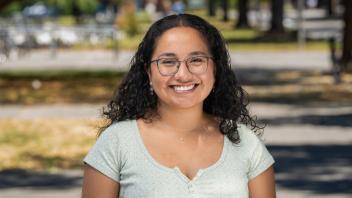 A person with long, curly hair and glasses is smiling outdoors. They are wearing a light green shirt and small earrings, and the background shows a blurred outdoor setting with trees and a pathway.