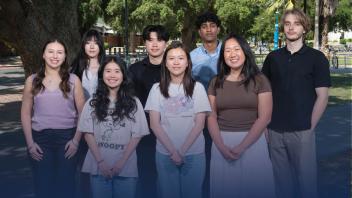 Group of eight students stand outdoors in sunlight, smiling at the camera with trees and bikes in the background.