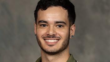 A person with facial hair in a brown shirt standing in front of a blurred background