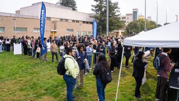 A large group of attendees lines up outside on a grassy lawn, bordered by buildings and flag banners that read "Information." People are casually dressed, and a white canopy tent is visible on the right.
