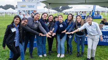 A group of smiling individuals under a tent poses for a photo with their hands outstretched in a unified gesture. Behind them is a UC Davis Health School of Medicine banner and rows of white tents in the background.
