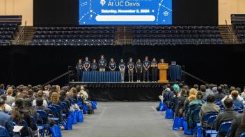 A stage at a UC Davis auditorium features a group of students in matching blue shirts addressing an audience. A large screen above them displays event details, including the title "Pre-Health Conference at UC Davis."