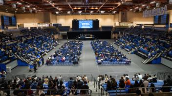 A wide-angle view of a UC Davis auditorium during a conference. Rows of attendees sit facing the stage, and a large screen displays event details. Blue conference bags are placed on seats throughout the audience.