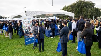 An outdoor event at UC Davis features attendees interacting at informational booths under large white tents. A UC Davis Health School of Medicine banner stands prominently in the foreground.