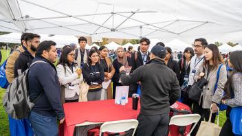 A group of attendees gathers around a red table under a white tent as a presenter gestures toward materials on the table. The attendees appear engaged and are wearing conference badges.