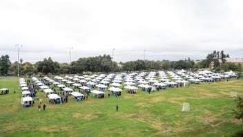 A wide aerial view of a large outdoor event, featuring rows of white canopy tents set up on a grassy field. Attendees can be seen walking between the tents, surrounded by trees and tall light poles in the background.