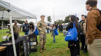 A U.S. Army recruiter, dressed in military fatigues, speaks to a group of attendees at an informational booth under a white canopy tent. The booth features Army materials and visuals, while attendees listen attentively, holding event bags.