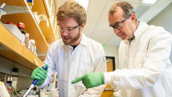Two researchers in lab coats and standing in a laboratory setting examining an object on a table.