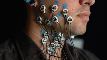 Close-up of a participant’s face and neck with multiple electrodes attached using blue medical tape. The electrodes are connected to thin black wires, leading to a monitoring device, highlighting a scientific study on muscle or nerve activity.